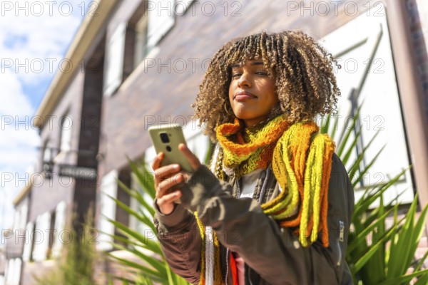 Latin young woman using phone standing in a residential street wearing warm clothes and scarf in a sunny day