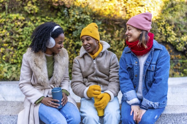 Three cheerful young friends are sitting on a bench in a city park during winter, holding reusable cups and enjoying a conversation