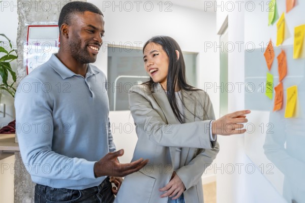 Two smiling businesspeople discussing work related tasks pointing at sticky notes on whiteboard in modern coworking office
