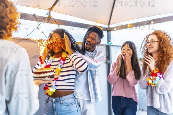 Multiracial young group of friends at terrace covering the eyes of a woman to surprise her with birthday cake