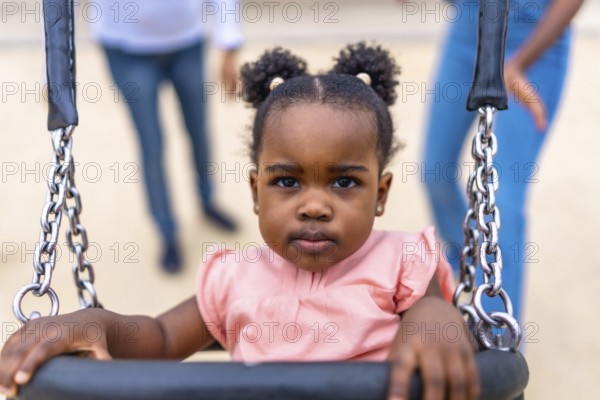 Cute african baby girl sitting on swing in a public park next to her family