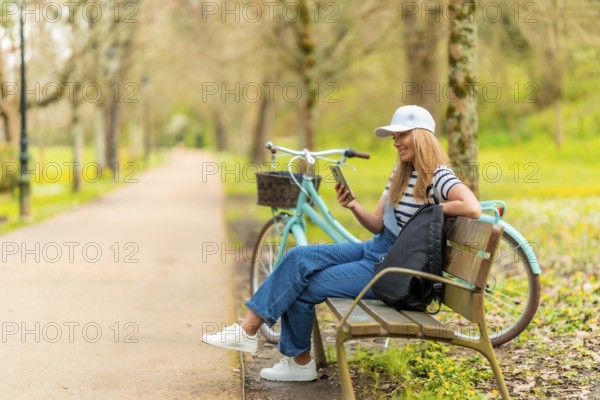 Blonde female tourist wearing casual clothing, using smartphone and resting on park bench with bicycle next to her
