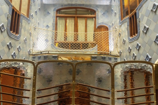 Light filtering through textured glass panels illuminates the unique blue tiled walls and wooden balcony of casa batllo, showcasing antoni gaudi's architectural brilliance