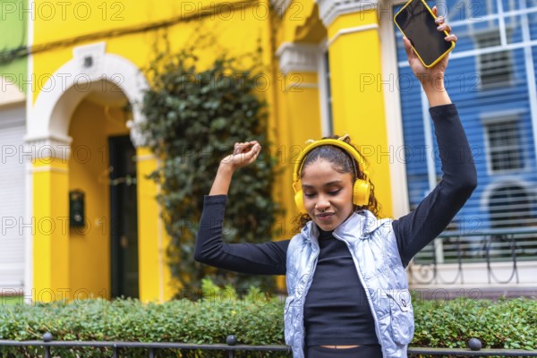 Happy african american woman dancing raising arms while listening to music with headphones and mobile phone in the city in a colorful street