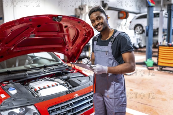 Smiling mechanic taking notes on clipboard while inspecting engine of red car with open hood in a professional workshop
