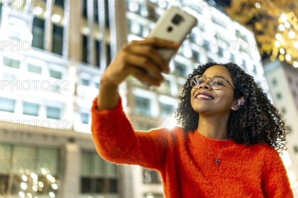Smiling woman capturing a selfie with her mobile phone in a vibrant urban city, surrounded by festive christmas lights at night