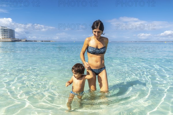 Mother and son holding hands and wading in the crystal clear turquoise waters of estanys beach in mallorca, spain, enjoying summer vacation