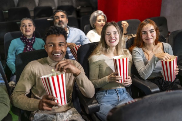 Multi ethnic group of friends enjoying a film at the movie theater, sharing laughter and munching on popcorn in a cozy auditorium setting