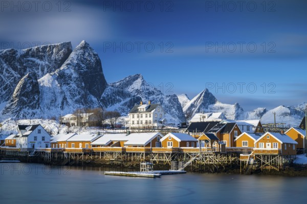 Traditional yellow rorbuer huts on the island of Sakrisøy, snow-capped mountains in the background, Sakrisoy, Reine, Lofoten, Norway, Europe