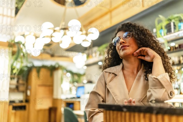 Elegant latin beauty woman with curly hair waiting hand on chin in a modern cafeteria