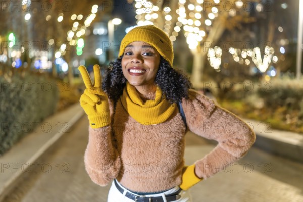 Smiling woman wearing yellow winter clothes showing victory sign while enjoying christmas lights in a city at night