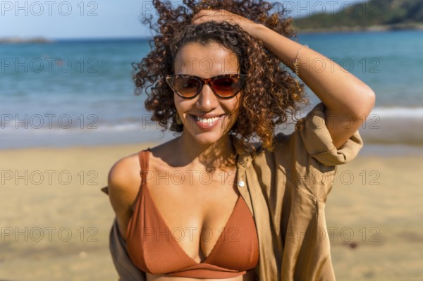 Sensual woman in bikini and hands on hair smiling at camera wearing sunglasses at beach