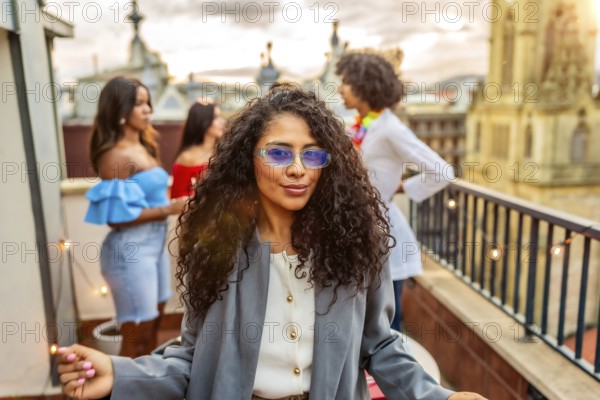 Cool woman in sunglasses smiling while friends partying on the background during rooftop evening party
