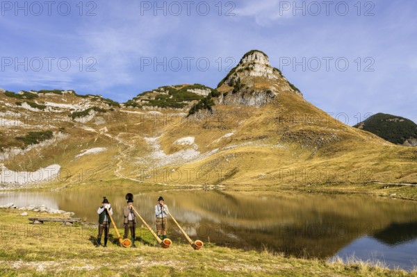 Three men play the alphorn at Lake Augstsee on Mount Loser. The Austrian alphorn trio Klangholz. Mount Atterkogel in the background. Autumn, good weather, blue sky. Reflection. Altaussee, Bad Aussee, Ausseer Land, Totes Gebirge, Styria, Upper Austria, Austria, Europe