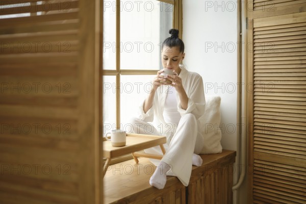 Pretty woman enjoys a peaceful morning, sipping coffee while sitting on windowsill, basking in the natural light