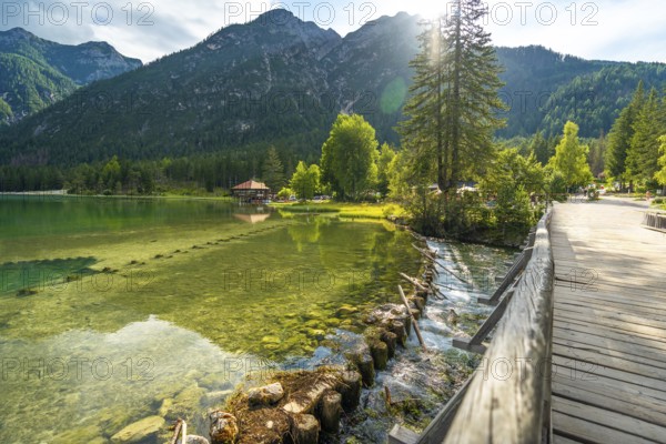 Sunlight illuminating the crystal clear waters of lake dobbiaco, with a wooden bridge and the dolomites in the background on a summer day