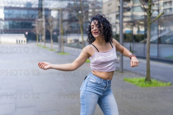 Curly haired hispanic woman dancing in urban city street with modern buildings in the background, wearing casual clothes