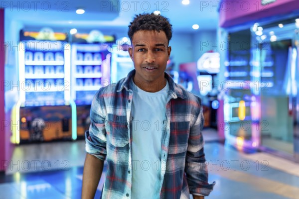 Young man smiles while standing in a lively arcade, surrounded by colorful claw machines and other games, enjoying his weekend leisure time