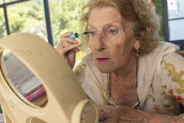 Senior woman applying mascara while gazing into a mirror, preparing for an outing. Embracing her beauty routine, she enjoys a moment of self-care and confidence indoors