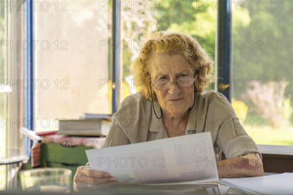 Focused elderly woman with glasses reviewing paperwork, possibly bills or medical records, sitting at her desk in a sunlit room with a view of greenery through the window