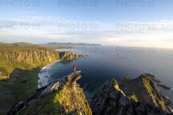 Evening mood, mountaineer standing on a rocky outcrop on the summit of Måtind, view of coastal landscape, Høyvika beach and sea, Måtinden, Andøya island, Vesterålen, Nordland Norway