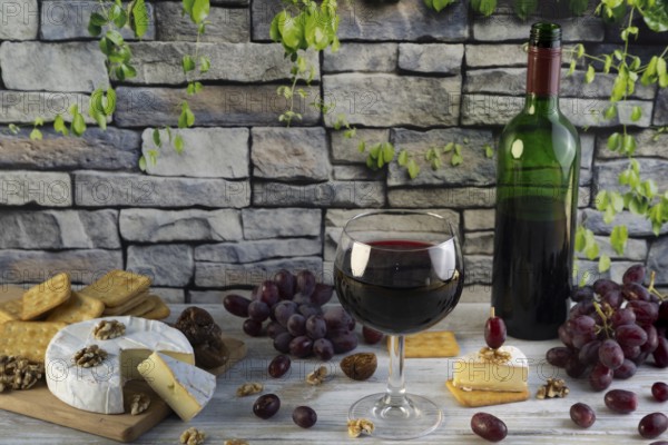 Wine glass and bottle of red wine surrounded by cheese, grapes, nuts and crackers in front of a rustic stone wall