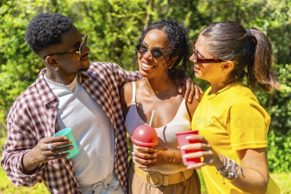 Three young multi ethnic friends enjoying a sunny day outdoors, holding drinks and smiling