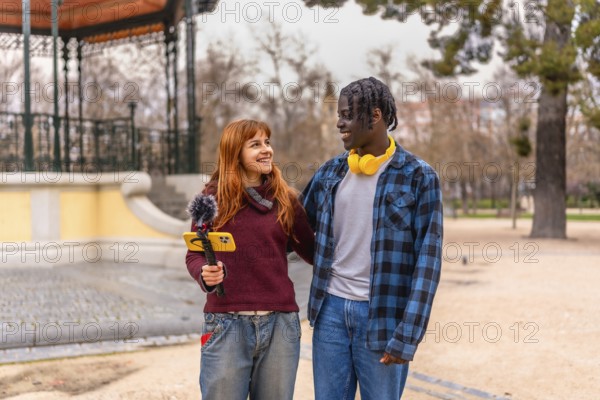 Two young content creators walking and talking in a park, recording audio with a mobile phone and microphone