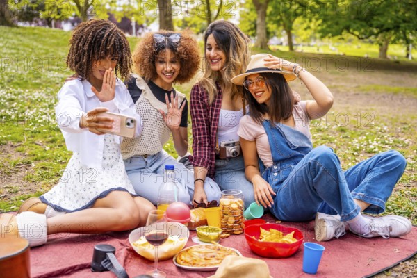 Four cheerful young women enjoying a picnic in a park, taking a joyful selfie with a smartphone while surrounded by nature and laughter