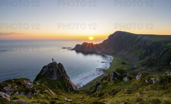 Climber on rocks in front of picturesque landscape, view of coastal landscape at sunset, cliffs and summit of Måtind or Måtinden, Høyvika beach and sea, Andøya island, Vesterålen, Nordland Norway