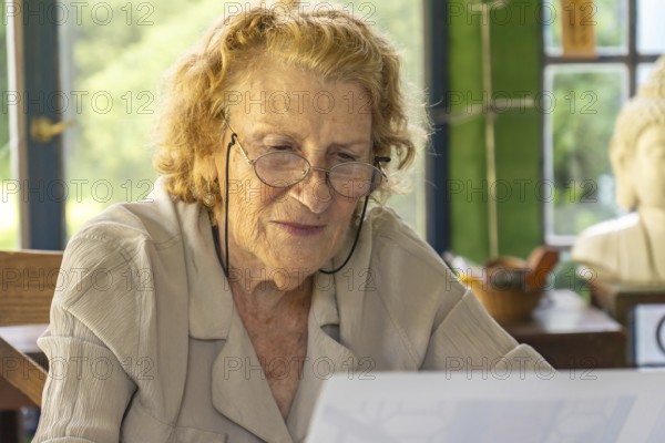 Elderly woman wearing glasses is sitting at a table, carefully reading documents with a focused expression on her face, possibly enjoying a peaceful moment at her home or in her studio