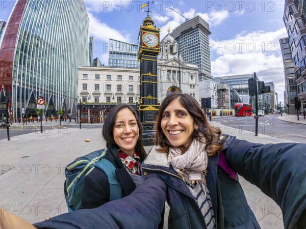 Two happy tourists taking a selfie in london near a historical clock tower, with modern buildings and red bus in the background