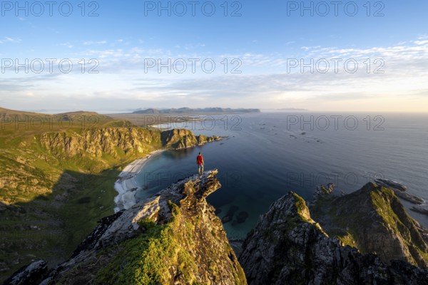 Evening mood, mountaineer standing on a rocky outcrop on the summit of Måtind, view of coastal landscape, Høyvika beach and sea, Måtinden, Andøya island, Vesterålen, Nordland Norway