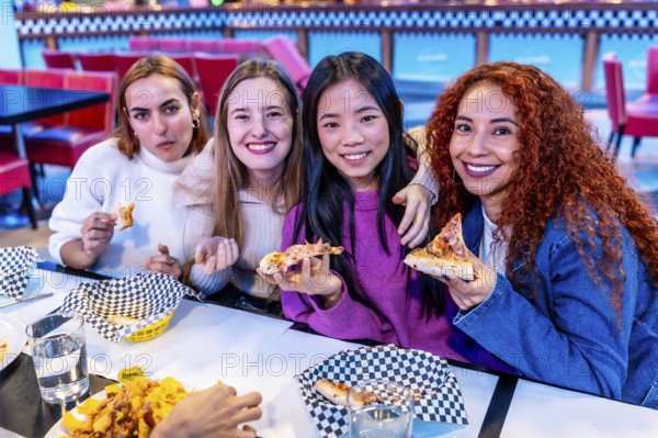 Four diverse young women enjoying a meal together at a restaurant, holding slices of pizza and smiling