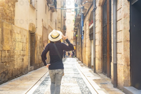 Female tourist wearing a straw hat walking down a narrow cobblestone street in the historic city center of tarragona, spain, enjoying a sunny day of sightseeing