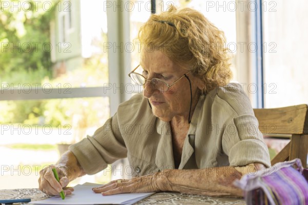 Concentrated elderly woman wearing glasses drawing with green colored pencil on sheet of paper while sitting at wooden table in light room near window at daytime