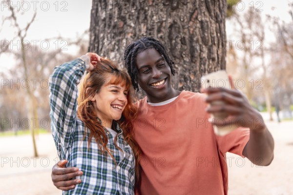 Young multiracial couple embracing and taking a selfie with a smartphone in a park, enjoying a beautiful day together