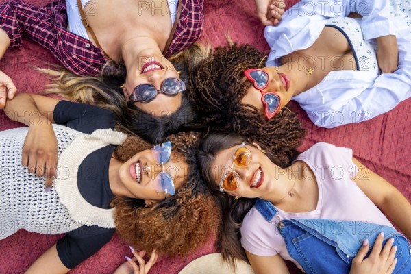 Four diverse young women lying together on a vibrant red blanket, smiling brightly at the camera while enjoying a sunny day outdoors