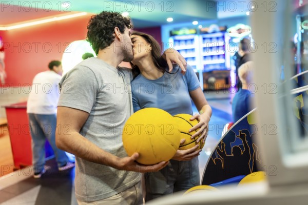 Romantic couple kissing while enjoying basketball and bowling in a vibrant entertainment center, surrounded by friends and arcade games
