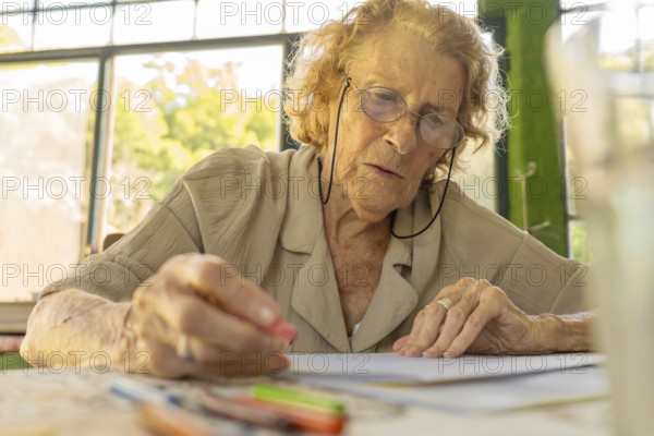 Concentrated elderly woman wearing glasses drawing with colored pencils at home, enjoying her hobby and expressing creativity in a peaceful and comfortable environment