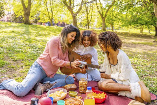 Three friends enjoying a picnic in a park, taking photos with a vintage camera, playing guitar, and sharing food and drinks on a sunny day