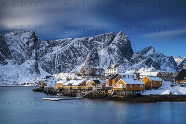 Traditional yellow rorbuer huts on the island of Sakrisøy, snow-capped mountains in the background, Sakrisoy, Reine, Lofoten, Norway, Europe
