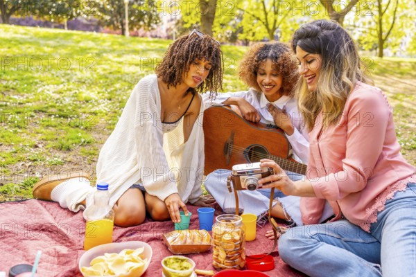 Three female friends enjoying a picnic in a park, taking pictures with a vintage camera, eating snacks, and playing guitar