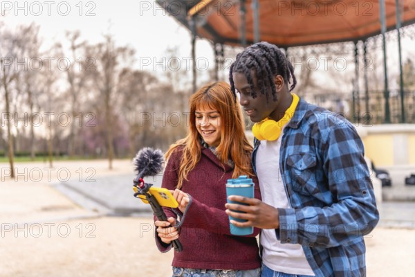 Two young content creators are recording a video in a park, using a smartphone with a microphone and reviewing the footage