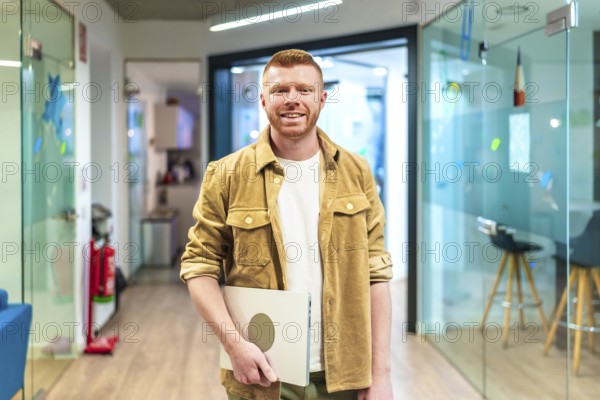 Young professional walking in a modern office corridor, holding a laptop and smiling, showcasing confidence and success in a dynamic work environment