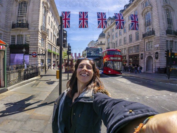 Young woman taking a selfie in london with union jack flags and a red double decker bus in the background, enjoying a sunny day