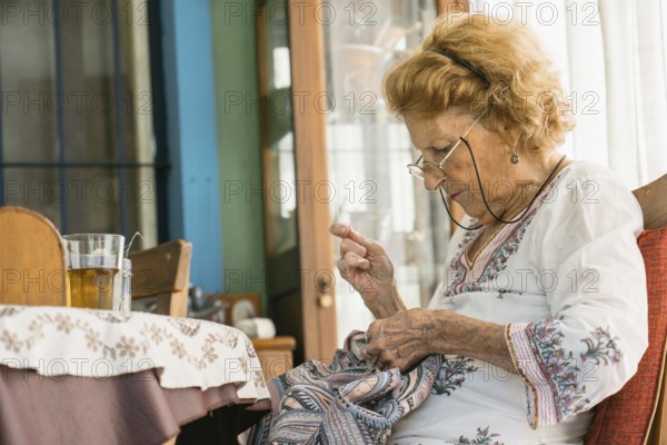 Senior woman enjoying retirement while sewing vibrant fabric, sitting comfortably beside a table with a cup of herbal tea, surrounded by a cozy and inviting home atmosphere