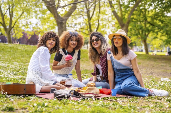 Four diverse female friends enjoying a picnic in a park, sharing food, drinks, and good times on a sunny day