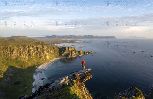 Evening mood, mountaineer standing on a rocky outcrop on the summit of Måtind, view of coastal landscape, Høyvika beach and sea, Måtinden, Andøya island, Vesterålen, Nordland Norway