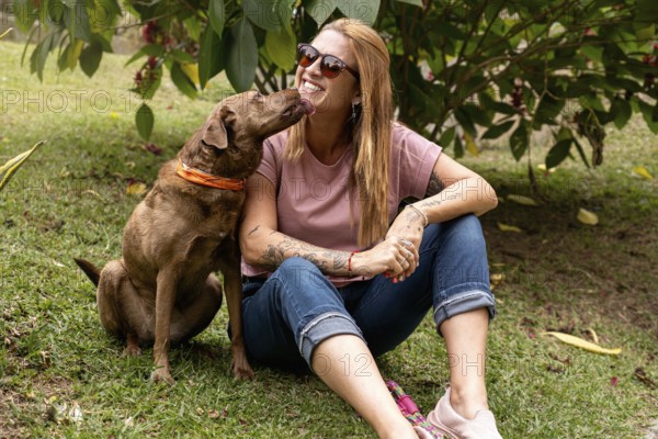 Woman in her 40s wearing sunglasses with a joyful expression receives a lick from her pet while they both sit on the grass, enjoying a sunny day in the park